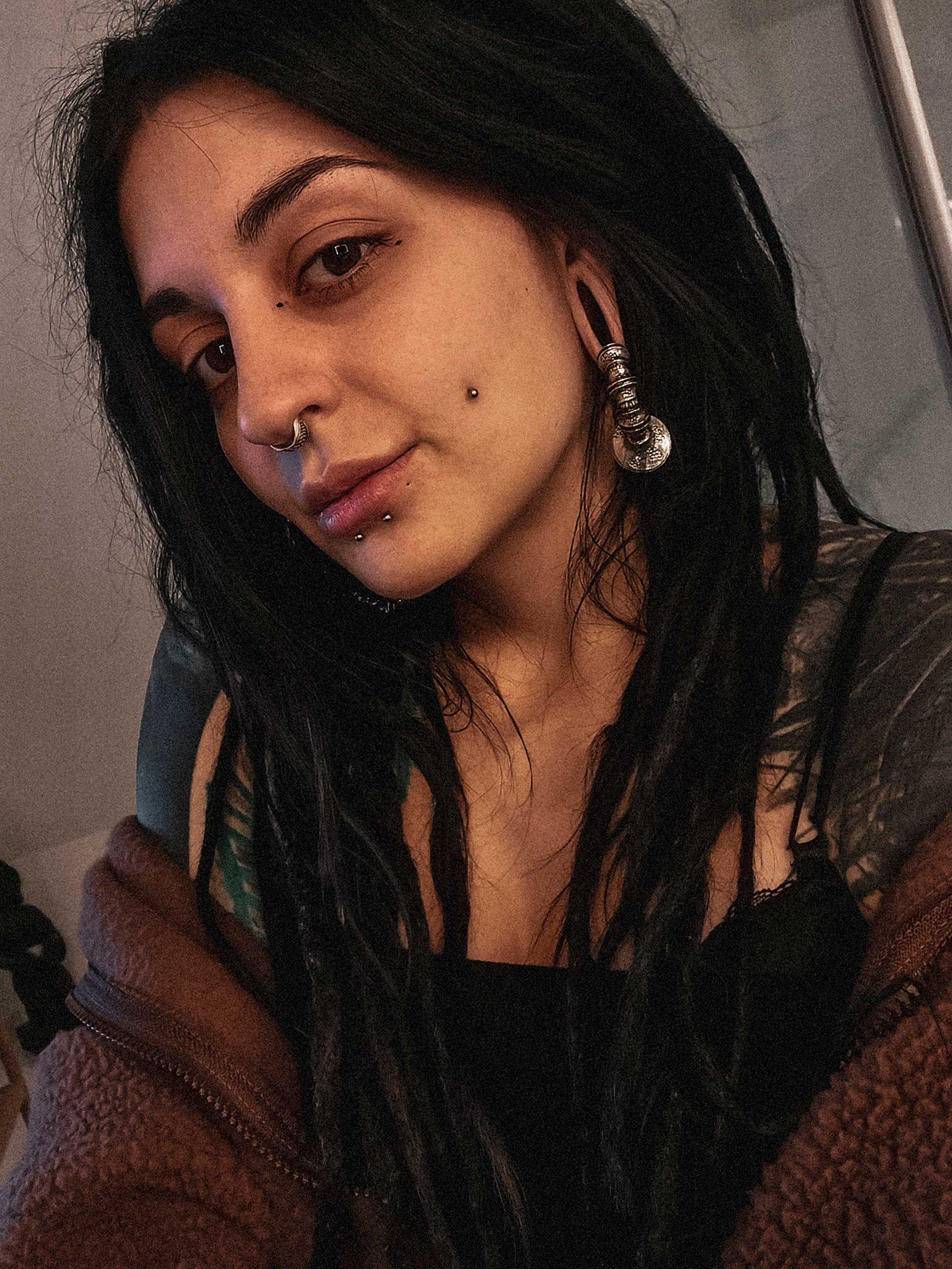 A woman with long, dark dreadlocks and noticeable earrings, including a large spiral gauge, poses for a selfie. Donning post-apocalyptic body jewelry and the Darma Biomechanical Clicker Hoop Septum Nose Ring in Silver by Kaya Movement, she showcases multiple facial piercings: a unisex 16 gauge nose piercing, lip piercing, and a dermal on her cheek. She wears a brown jacket and black top.